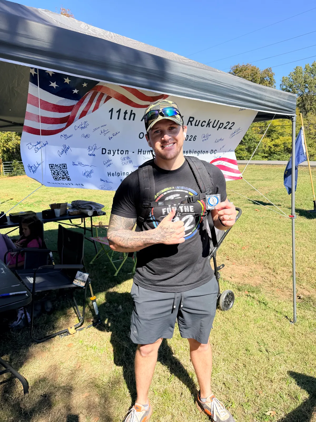 Cody Hitson in a For The 22 shirt giving a thumbs up and holding the 22-mile coin under the RuckUp22 event tent