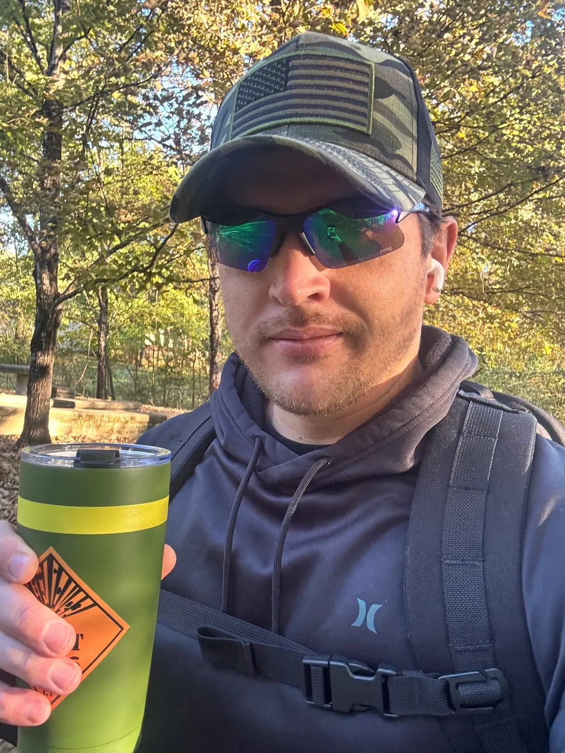 Cody Hitson in a camo flag cap and sunglasses holding up a green coffee tumbler on a wooded trail in the morning light