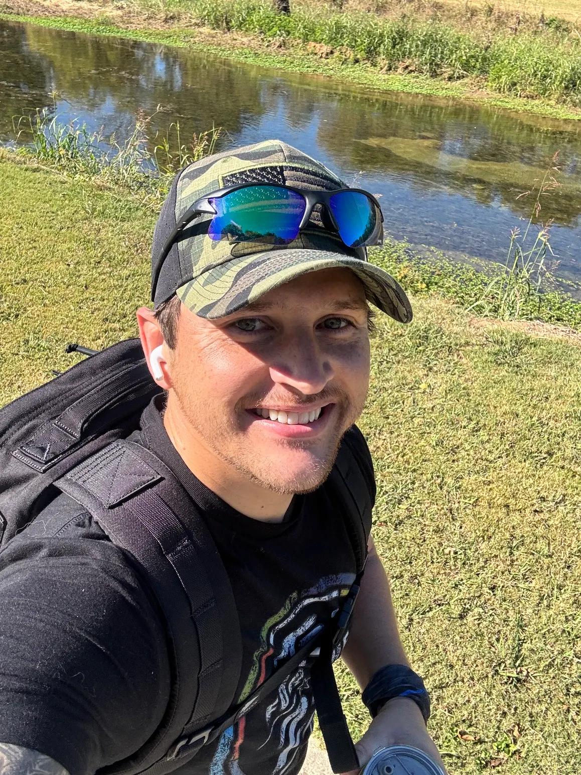 Cody Hitson smiling in a selfie with a ruck on his back, a pond and grass behind him